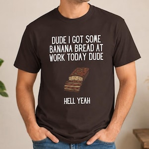 May include: A dark brown short-sleeve t-shirt featuring white text that reads DUDE I GOT SOME BANANA BREAD AT WORK TODAY DUDE and HELL YEAH, with a graphic illustration of a sliced loaf of banana bread positioned between the lines of text.