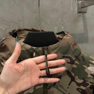 Pu&ograve; includere: Giacca mimetica con collo alto e chiusura a zip. Presenta una combinazione di colori marrone, verde e beige. Il logo Arc'teryx &egrave; visibile in basso a destra.