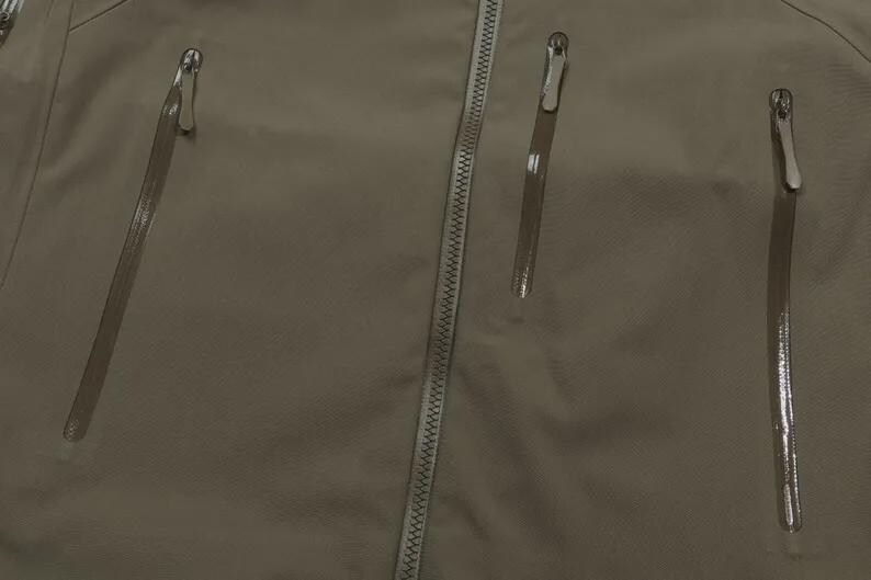 May include: Close-up of a khaki jacket featuring a central zip and three vertical zipped pockets. The zips have silver pulls. The fabric looks smooth and water-resistant, suitable for outdoor wear.