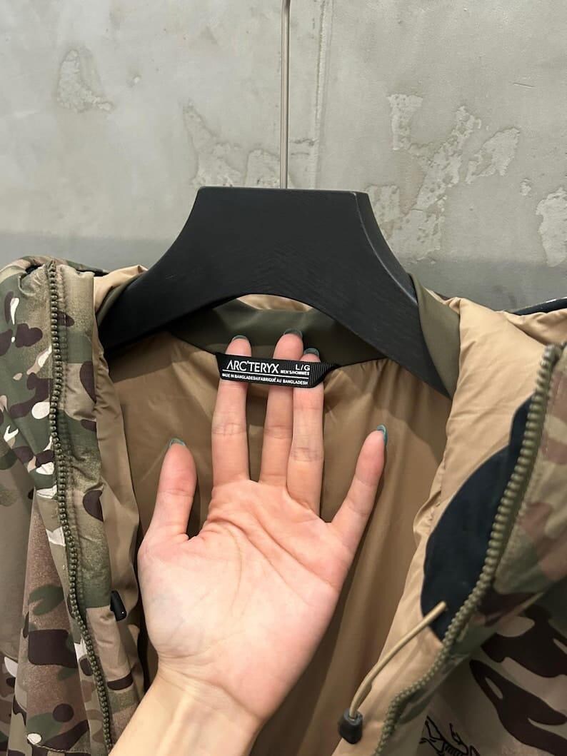 Pu&ograve; includere: Giacca mimetica con un motivo nei toni del verde, marrone e beige, appesa a una gruccia nera. L'etichetta all'interno del colletto mostra il marchio "ARC'TERYX".