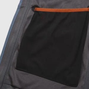 May include: Detail of a grey jacket interior, showing a large black pocket with a brown trim. The jacket has blue edging and a zip detail.