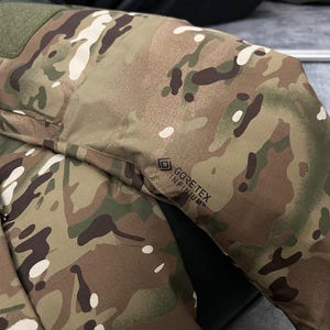 Pu&ograve; includere: Primo piano di una giacca mimetica con motivi verdi, marroni e bianchi. La giacca presenta una toppa verde sulla spalla e il logo "GORE-TEX INFINIUM".