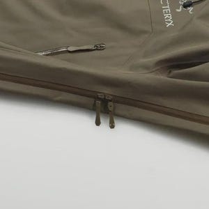 May include: A close-up of a khaki-coloured jacket featuring a zip closure and a side pocket. The jacket has a minimalist design with the brand name "Arc'teryx" visible. The material appears to be water-resistant.