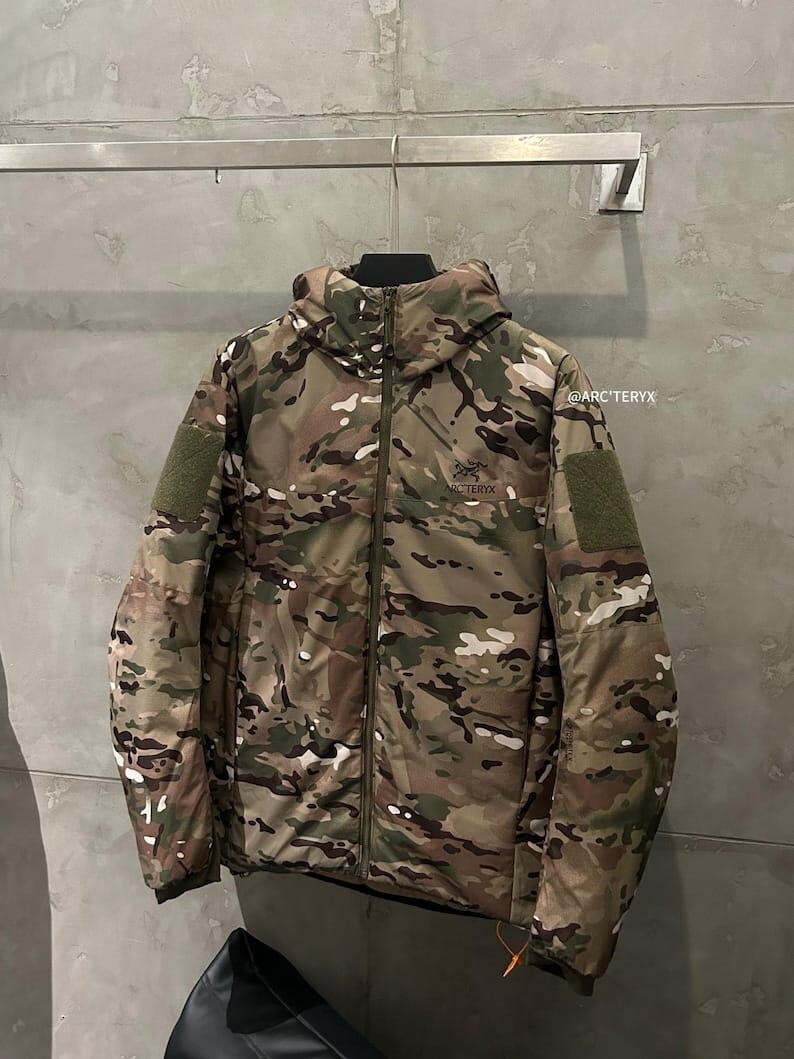 Pu&ograve; includere: Giacca con motivo mimetico con cappuccio, chiusura a zip e due toppe verdi sulle maniche. La giacca, con il logo Arc'teryx, &egrave; appesa a una barra di metallo contro un muro grigio.