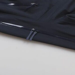 May include: Close-up of a navy blue garment with a shiny, waterproof finish. The image shows two zippers and a horizontal seam with a reflective strip. The fabric is folded, creating shadows and texture.