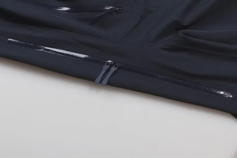May include: Close-up of a navy blue garment with a shiny, waterproof finish. The image shows two zippers and a horizontal seam with a reflective strip. The fabric is folded, creating shadows and texture.