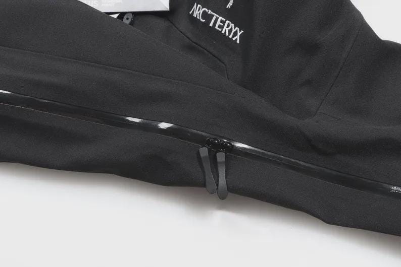 May include: A close-up of a black jacket featuring a zipper and the brand name "ARC'TERYX" in white. The jacket is made of a smooth, matte material. The zipper has two pulls.