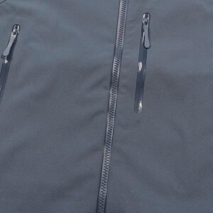 May include: A close-up view of a slate blue jacket, featuring three vertical zipped pockets and a central zip. The zips have silver pulls, contrasting with the smooth, matte fabric. The jacket appears to be made from a lightweight, water-resistant material.