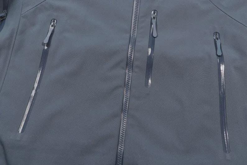 May include: A close-up view of a slate blue jacket, featuring three vertical zipped pockets and a central zip. The zips have silver pulls, contrasting with the smooth, matte fabric. The jacket appears to be made from a lightweight, water-resistant material.