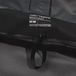 May include: A close-up of a black Arc'teryx label, with the text "MEN'S/HOMMES" and size "M/M". The label also includes the text "MADE IN CHINA / FABRIQU&Eacute; EN CHINA". A black loop is attached to the label.