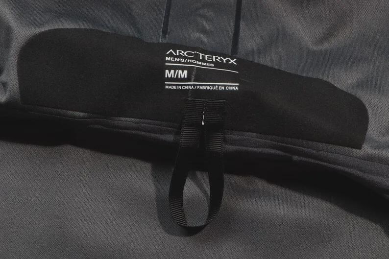 May include: A close-up of a black Arc'teryx label, with the text "MEN'S/HOMMES" and size "M/M". The label also includes the text "MADE IN CHINA / FABRIQU&Eacute; EN CHINA". A black loop is attached to the label.