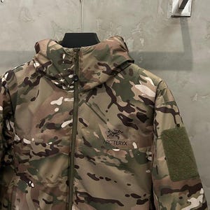 Pu&ograve; includere: Giacca mimetica con cappuccio e chiusura a zip. La giacca presenta il logo "ARC'TERYX" sul petto e una toppa in velcro sulla manica. La giacca &egrave; appesa a un gancio di metallo. Il motivo include tonalit&agrave; di verde, marrone e beige.