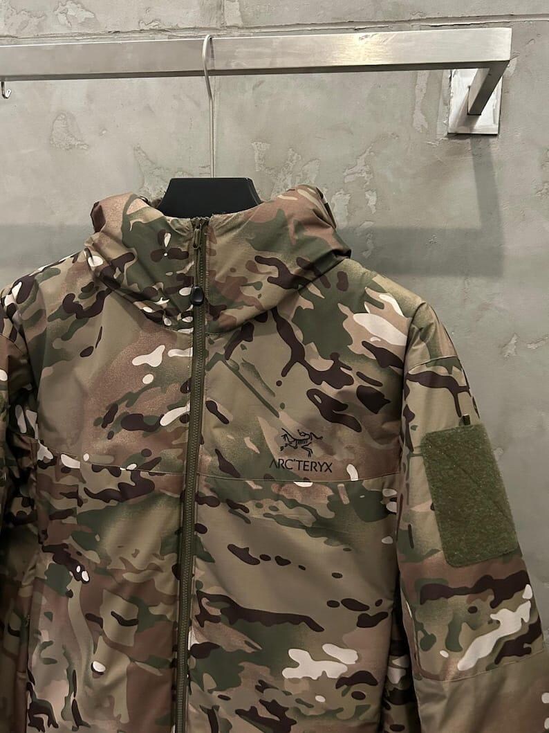 Pu&ograve; includere: Giacca mimetica con cappuccio e chiusura a zip. La giacca presenta il logo "ARC'TERYX" sul petto e una toppa in velcro sulla manica. La giacca &egrave; appesa a un gancio di metallo. Il motivo include tonalit&agrave; di verde, marrone e beige.