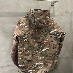 Pu&ograve; includere: Giacca mimetica con cappuccio nei toni del marrone, verde e bianco appesa a un supporto metallico. La giacca presenta un collo alto e una cerniera a tutta lunghezza. Il marchio "@ARCTERYX" &egrave; visibile in alto a sinistra.