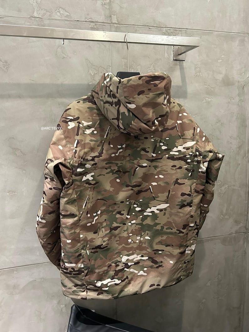 Pu&ograve; includere: Giacca mimetica con cappuccio nei toni del marrone, verde e bianco appesa a un supporto metallico. La giacca presenta un collo alto e una cerniera a tutta lunghezza. Il marchio "@ARCTERYX" &egrave; visibile in alto a sinistra.