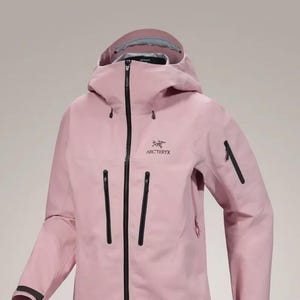 May include: A pale pink hooded jacket with a full-length black zip and several black zipped pockets. The jacket displays the "Arc'teryx" logo on the chest and black detailing on the sleeves.