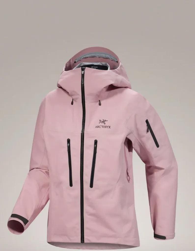 May include: A pale pink hooded jacket with a full-length black zip and several black zipped pockets. The jacket displays the "Arc'teryx" logo on the chest and black detailing on the sleeves.
