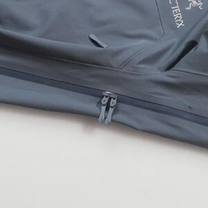 May include: A close-up of a slate-grey jacket with a zip. The jacket displays the Arc'teryx logo and Gore-Tex branding. The fabric looks smooth and water-resistant, with a slight sheen.