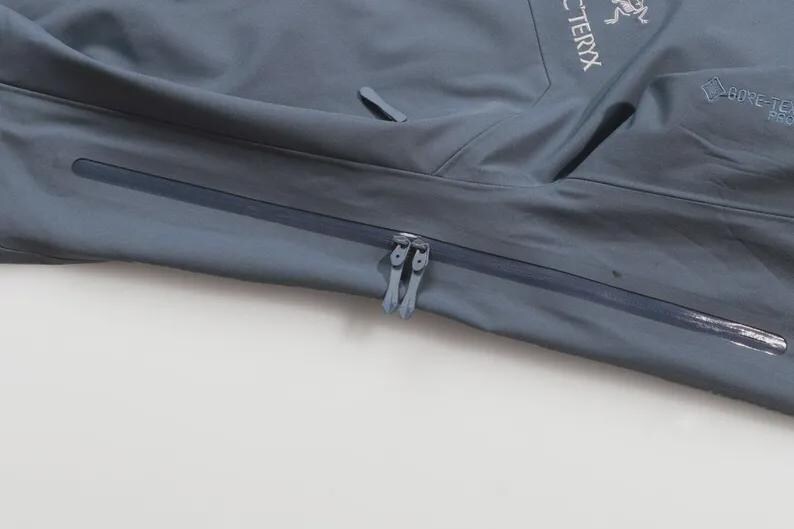 May include: A close-up of a slate-grey jacket with a zip. The jacket displays the Arc'teryx logo and Gore-Tex branding. The fabric looks smooth and water-resistant, with a slight sheen.