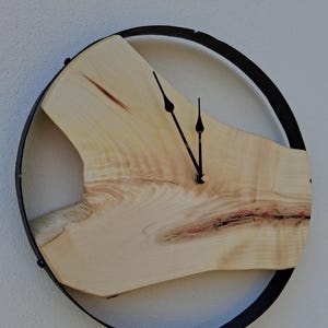 Könnte beinhalten: Eine Wanduhr bestehend aus einer hellen Naturholzscheibe mit sichtbarer Maserung und einem dunklen Riss, die in einem dünnen, kreisförmigen schwarzen Metallrahmen hängt. Die Uhr hat zwei schmale schwarze Zeiger in der Mitte des Holzes.