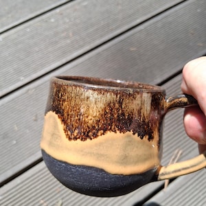Könnte beinhalten: Ein Keramikbecher mit einem dreifarbigen Glasurdesign. Der obere Teil hat eine dunkelbraune, gesprenkelte Oberfläche, das mittlere Band ist glatt und hellbraun, und der Boden ist in mattem, texturiertem Schwarz gehalten. Der Becher hat einen Henkel.