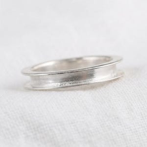 Könnte beinhalten: Ein silberner Ring mit glatter, polierter Oberfläche und einer ausgeprägten Rille, die mittig entlang des Bandes verläuft. Der Ring weist ein leicht konkaves Profil zwischen zwei erhöhten Kanten auf, was für ein schlichtes metallisches Design sorgt.