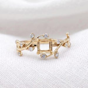 May include: A gold ring featuring a central square setting designed to hold a gemstone. The band is crafted in a vine-like motif with small, clear, round stones embedded along the branches, creating a delicate and intricate metal structure.