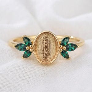 May include: A gold-toned ring featuring a central oval setting with a textured surface, flanked on each side by three marquise-cut dark green stones arranged in a leaf-like pattern. The band is thin and polished, showcasing a metallic finish.