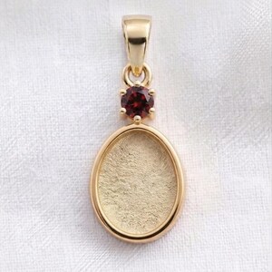 May include: A gold pendant featuring a smooth, oval-shaped setting with a textured, matte gold interior. Above the oval frame sits a single, round, deep red garnet gemstone held by four prongs, connected to a polished gold bail at the very top.