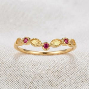 May include: A gold band ring featuring a curved, chevron-like shape. The ring is adorned with three small, round, vibrant pink gemstones set within textured, marquise-shaped gold settings that alternate with smooth, polished gold sections.