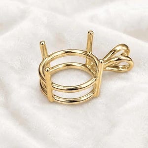 May include: A gold-toned metal pendant setting featuring a circular frame with four vertical prongs designed to hold a gemstone. The piece includes a decorative, looped bail attached to the side for threading onto a chain or necklace cord.