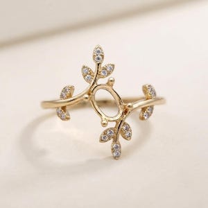 Könnte beinhalten: Ein goldener Ring mit einer zentralen, ovalen, offenen Fassung, die von zwei symmetrischen, rankenartigen Zweigen flankiert wird. Jeder Zweig ist mit kleinen, klaren, funkelnden Steinen besetzt, die in die Metallblätter eingelassen sind.
