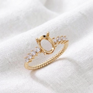 May include: A gold ring featuring a central oval-shaped prong setting designed to hold a gemstone. The band has a twisted rope texture and is accented on both sides of the center setting with three small, round, clear, sparkling stones.
