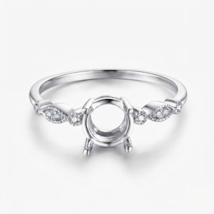 Puede incluir: Un engaste de anillo en tono plateado con una montura central de cuatro garras diseñada para sostener una piedra. La banda está decorada con pequeñas piedras redondas y transparentes incrustadas en engastes metálicos en forma de marquesa.