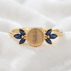 May include: A gold ring featuring a central circular setting with a textured surface, flanked on each side by three marquise-cut deep blue stones arranged in a fan-like pattern. The band is thin and polished, creating a balanced and symmetrical design.