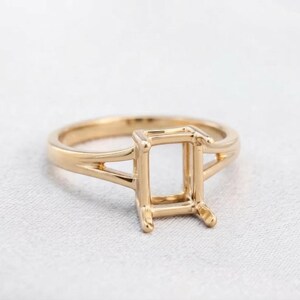 Puede incluir: Un anillo de oro pulido con un engaste rectangular de garras diseñado para sostener una piedra preciosa. La banda presenta un diseño de brazo dividido que se estrecha hacia el engaste central, creando un aspecto limpio y estructurado.