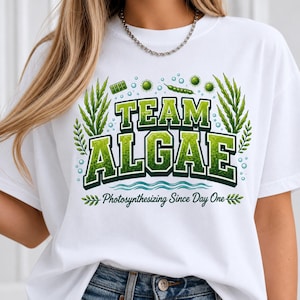 Peut inclure: Un t-shirt blanc arborant un motif avec le texte "TEAM ALGAE" et "Photosynthesizing Since Day One" en lettres vertes. Le design comprend des illustrations d'algues vertes, de bulles et d'organismes microscopiques dans diverses nuances de vert.