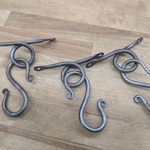Könnte beinhalten: Drei handgeschmiedete, graue Metall-S-Haken. Jeder Haken hat ein geschlungenes Design mit einer S-förmigen Kurve und einer flachen, rechteckigen Oberseite mit zwei Löchern zur Befestigung. Die Haken sind zum Aufhängen von Gegenständen bestimmt.