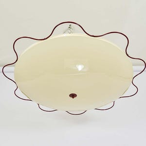 Puede incluir: Una lámpara de techo con una pantalla circular lisa de color crema. La pantalla está rodeada por un marco de metal fino de color rojo oscuro con un borde ondulado y festoneado. Un pequeño pomo circular rojo oscuro está centrado en la base.