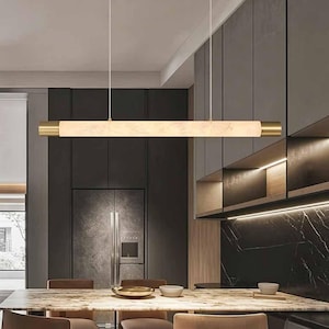 May include: A linear pendant light fixture featuring a long, cylindrical, cream-colored marble-patterned shade suspended by two thin white cords. The ends of the cylinder are capped with brushed brass metal accents, creating a sleek horizontal profile.