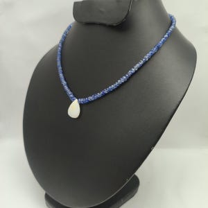 May include: A blue beaded necklace with a pear-shaped, white pendant. The necklace is displayed on a black jewelry stand against a white background. The beads appear to be small and evenly spaced, creating a delicate design.