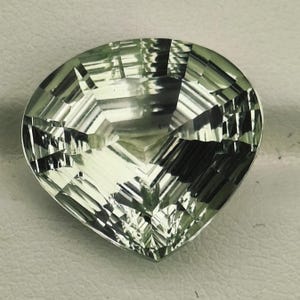 Könnte beinhalten: Ein herzförmiger Edelstein mit blassgrünem Farbton und facettierter Oberfläche. Der Stein weist zahlreiche geometrische Schliffe auf, die ein reflektierendes, mehrschichtiges Erscheinungsbild mit Hell-Dunkel-Kontrast auf dem polierten Körper erzeugen.