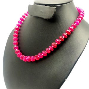 May include: A bright pink beaded necklace on a black jewellery stand. The necklace is made of small, round beads in shades of pink and red. The beads are strung together to form a complete circle, suitable for wearing.