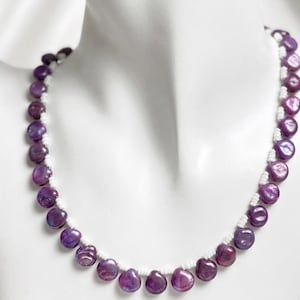 Puede incluir: Un collar con grandes cuentas redondas de color púrpura con un brillo nacarado. Las cuentas están ensartadas en un cordón blanco, creando un contraste llamativo. El collar se exhibe en un maniquí blanco.