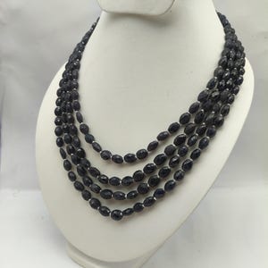 May include: A multi-strand necklace with dark blue, faceted beads. The necklace is displayed on a white bust, showcasing the layered design and the color of the beads. The beads have a slightly oval shape.