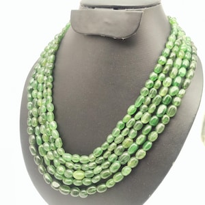 May include: A multi-strand necklace featuring oval-shaped green beads. The necklace is displayed on a black jewelry bust. The beads have a polished finish and vary in shades of green.