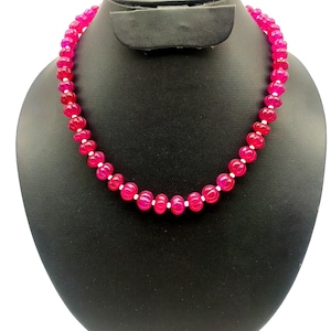 Op de afbeelding: Een felroze kralenketting op een zwarte sieradenstandaard. De ketting heeft ronde, gepolijste kralen met kleine witte tussenkralen. De kralen zijn diep, rijk roze van kleur, wat een opvallend effect creëert.