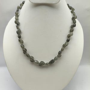 Puede incluir: Un collar compuesto por numerosas cuentas pequeñas de forma ovalada en un color gris translúcido con inclusiones oscuras y moteadas. Las cuentas están ensartadas en una sola hebra, creando un diseño circular uniforme y continuo.