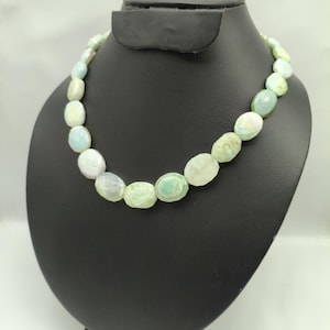 May include: A necklace featuring oval-shaped beads in shades of green, white, and pale blue. The beads are strung together, creating a statement piece. The necklace is displayed on a black jewelry bust against a white background.