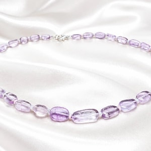 Op de afbeelding: Een ketting met ovale amethist kralen in verschillende tinten paars en lavendel. De kralen zijn geregen met een zilveren sluiting, waardoor een delicaat en elegant sieraad ontstaat.
